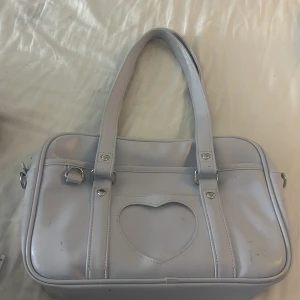 Ljuslila axelväska med hjärtdetalj - Säljer en söt ljuslila axelväska i med ett hjärta på framsidan. Väskan har dubbla handtag och silverfärgade detaljer. Perfekt för att ge en gullig touch till din outfit. Köpt för 500kr