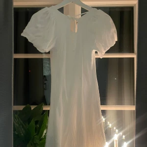 Vit klänning med puffärmar - Säljer en stilren vit klänning med korta puffärmar och rund hals. Klänningen har en ribbad struktur och en liten knytning i nacken. Perfekt för sommarens alla tillfällen!