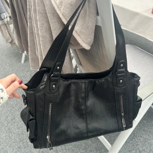 Svart axelväska  - Snygg svart axelväska i syntetmaterial med flera praktiska dragkedjor och fickor. Perfekt för att hålla ordning på dina saker. Väskan har robusta handtag och en stilren design. Helt ny för råka beställa två. Nypris 600, mitt pris 300. Går att förhandla vid snabb affär 