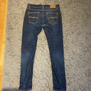 Mörkblå jeans från Nudie Jeans - Snygga mörkblå jeans från Nudie Jeans med klassisk femficksdesign och kontrastsömmar. De har en slim passform och är tillverkade i högkvalitativt denim. Perfekta för en stilren look. Har en liten slitning i mitten, men har fixats och syts ihop. Går att pruta vid snabb affär 