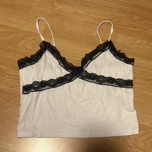 Vitt linne med spets - Säljer en söt vit linnetopp från FB Sister med svarta spetsdetaljer längs kanterna. Toppen har tunna axelband och en croppad passform. Storlek S💗