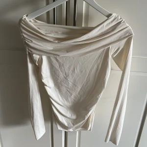 Vit offshoulder topp från Weekday - Säljer en oanvänd stilren krämvit offshoulder topp från Weekday. Toppen har långa ärmar och en elegant drapering över axlarna. Perfekt för en chic och modern look. Passar bra till både jeans och kjol. Orginal pris: 500