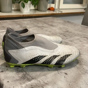 Adidas Predator fotbollsskor - Snygga Adidas Predator fotbollsskor i vitt och grått med svarta och limegröna detaljer. Skorna har en strömlinjeformad design utan snörning för en tight passform och optimal bollkontroll. Perfekta för fotbollsplanen med sina greppvänliga dobbar.  Jag har använt dom typ 5 gånger du behöver bara tvätta dom och jag köpte dom för 1300kr