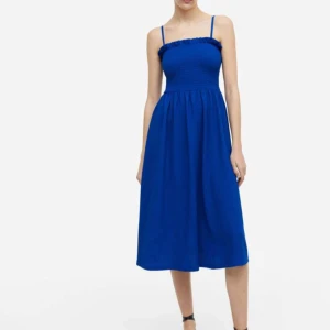 Blå klänning med smala axelband - Snygg blå klänning med smala axelband och smockad överdel. Perfekt för sommarens varma dagar. Klänningen har en luftig passform och når till knäna.