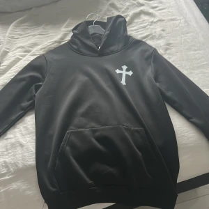 Svart hoodie med korsdetalj - Säljer en stilren svart hoodie med en vit korsdetalj på bröstet. Tröjan har en klassisk design med huva och en stor ficka framtill. Perfekt för en avslappnad look.