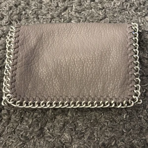 Tiamo väska - Snygg grå axelväska i skinn med coola kedjedetaljer runt kanterna. Väskan har en praktisk dragkedja på baksidan och en rymlig insida med extra fack. Perfekt för att ge din outfit en edgy touch! Endast lite skadad på baksida!