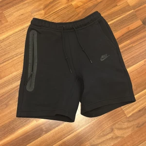 Svarta Nike tech shorts  - Snygga svarta shorts från Nike med snörning i midjan och en praktisk dragkedja på sidan. Perfekta för träning eller en avslappnad dag. Nike-loggan är diskret placerad på benet. Perfekt för sommaren