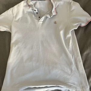 Moncler piké - Säljer en stilren vit pikétröja från Moncler med klassisk krage och knappar. Tröjan har en broderad Moncler-logga på bröstet och röd samt blå randdetalj på kragen. Perfekt för en avslappnad men ändå elegant look. Väldigt fint skick och passar till både festlig och vardags användning. Hör av er vid funderingar/ R.EsellingSWE😎