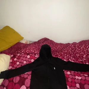 Svart hoodie från Ralph Lauren - Säljer en stilren svart hoodie från Ralph Lauren med det klassiska vita polo-logotypen på bröstet. Hoodien har dragkedja och är långärmad, perfekt för en casual look. Märkesetiketten är tydligt synlig inuti.