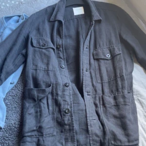 svart overshirt från a day’a march - Säljer en stilren svart overshirt med knappar framtill. Jackan har två bröstfickor med knappar och två stora fickor nedtill. Perfekt för lager-på-lager-stil och passar bra till både jeans och chinos.