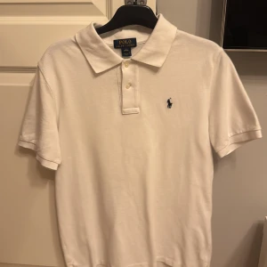 Vit pikétröja från Ralph Lauren - Säljer en klassisk vit pikétröja från Ralph Lauren med den ikoniska broderade loggan på bröstet. Tröjan har korta ärmar och en stilren krage med knappar. Perfekt för en avslappnad men ändå stilfull look.
