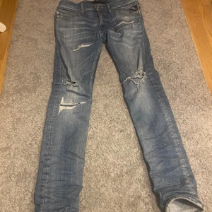 Blå jeans från Replay - Säljer ett par blå Replay Jondrill jeans med slitna detaljer och en snygg tvätt. De har en knappgylf och klassisk femficksdesign. Perfekta för en avslappnad stil.