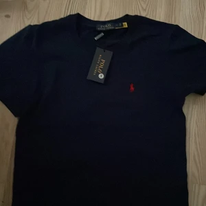 Mörkblå t-shirt från Polo Ralph Lauren - Snygg mörkblå t-shirt från Polo Ralph Lauren med det klassiska röda broderade logotypen på bröstet. Perfekt för en stilren och avslappnad look. T-shirten är kortärmad och gjord i mjukt material för extra komfort.