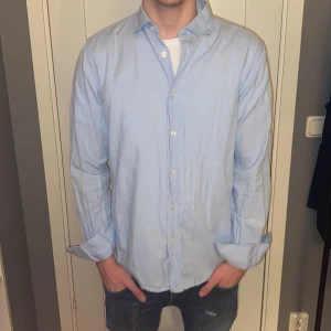 Ljusblå skjorta från J.Lindeberg - Snygg ljusblå skjorta från J.Lindeberg i herrstorlek S. Skjortan har en klassisk krage och knappar framtill. Perfekt för en stilren look.