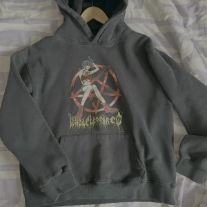 Playboicarti merch hoodie med anime-tryck - Säljer en grå hoodie med ett coolt anime-tryck på framsidan. Trycket föreställer en karaktär med blått hår och röda kläder framför en röd symbol. Perfekt för dig som gillar unik stil och anime-inspirerade plagg.