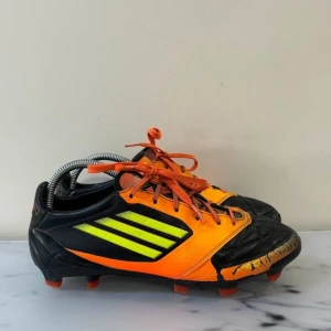 Svarta och orange fotbollsskor från Adidas - Säljer ett par coola fotbollsskor från Adidas i svart och orange med neongula detaljer. Skorna har snörning och är perfekta för fotbollsplanen. De har en sportig design med robust sula för bra grepp.