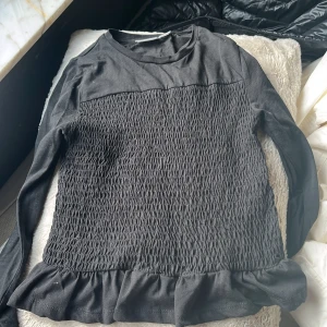 Svart långärmad topp med volang - Snygg svart långärmad topp med smockdetaljer på framsidan och volangkant nedtill. Perfekt för en stilren look. Passar bra till både jeans och kjol.