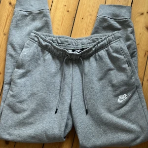 Grå mjukisbyxor från Nike - Säljer ett par grå mjukisbyxor från Nike med elastisk midja och snörning. Byxorna har ribbade muddar vid bensluten och en broderad Nike-logga på sidan. Bra skick! 