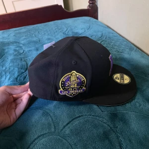 Svart keps med Los Angeles-broderi - Snygg svart keps från New Era med broderad Los Angeles-logga i guld och lila. Kepsen har en platt skärm och är i storlek 7 5/8 (60.6 cm). Perfekt för att visa ditt stöd för laget! Limited Edition.