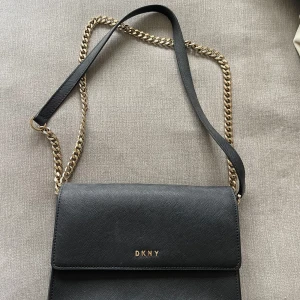 Svart axelväska från DKNY - Snygg svart axelväska från DKNY med guldfärgad kedja och logotyp framtill. Väskan har en stilren design och är perfekt för att bära dina nödvändigheter. Den har en elegant look som passar till många tillfällen. Nypris: ungefär 3500kr. 