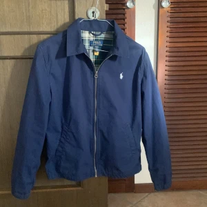 Blå jacka från Polo Ralph Lauren - Snygg blå overshirt från Polo Ralph Lauren med dragkedja och krage. Jackan har ett diskret broderat logotyp på bröstet och är perfekt för en stilren look. Den har långa ärmar och en bekväm passform.