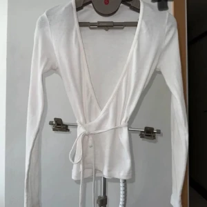 Vit omlottopp i bomull - Säljer en stilren vit omlottopp i bomull med långa ärmar. Toppen har en knytning i midjan som ger en smickrande passform. Perfekt för en elegant och avslappnad look.