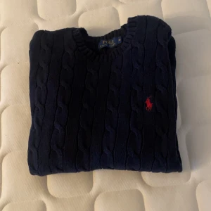 Mörkblå stickad tröja från Polo Ralph Lauren - Säljer en klassisk mörkblå stickad tröja från Polo Ralph Lauren med kabelstickat mönster. Tröjan har en rund halsringning och det ikoniska röda logotypbroderiet på bröstet. Perfekt för en stilren look. PASSAR storlek S !
