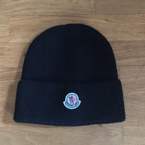 Svart stickad mössa från Moncler - Använd en gång. Snygg svart stickad mössa från Moncler med broderad logotyp framtill. Perfekt för kyliga dagar och ger en stilren look. Mössan är ribbad och har en klassisk design.