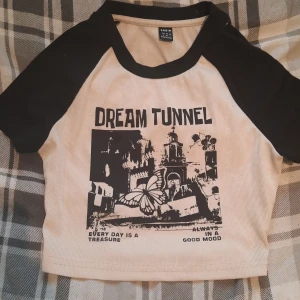 Beige och svart croppad topp - Säljer en cool croppad topp från Shein i beige med svarta ärmar och tryck framtill. Texten 'Dream Tunnel' och ett grafiskt motiv ger en unik look. Perfekt för en avslappnad stil.
