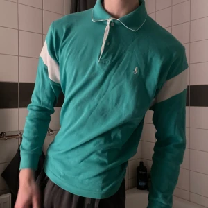 Turkos långärmad pikétröja - Snygg turkos långärmad pikétröja från Ralph Lauren med vita detaljer på ärmarna och kragen. Perfekt för en stilren och avslappnad look. Tröjan är en egen design, det vita på ärmarna är tillagd efter att tröjan är köpt vilket innebär att det inte finns någon likadan tröja. Riktigt fräsch och fin.