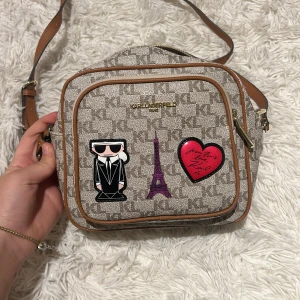 Beige axelväska från Karl Lagerfeld - Snygg beige axelväska från Karl Lagerfeld med coola detaljer som en Eiffel-torn och hjärta. Väskan har ett mönster med märkets logga och en praktisk dragkedja. Perfekt för att ge din outfit en touch av Paris.