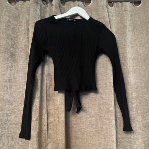 Svart ribbad topp med knyt - Snygg svart ribbad topp från SSTB i storlek S. Toppen har långa ärmar och en knytning i midjan som ger en fin detalj. Perfekt för en stilren look.