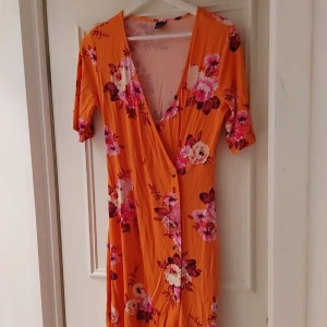 Orange blommig omlottklänning från Gina Tricot - Säljer en färgglad orange omlottklänning från Gina Tricot med vackert blommönster i rosa och vita nyanser. Klänningen har en v-ringad design och trekvartsärmar, perfekt för sommarens varma dagar. 🌸