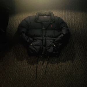 Marinblå dunjacka från Ralph Lauren - Säljer en snygg marinblå dunjacka från Ralph Lauren med röd broderad logga på bröstet. Jackan har dragkedja och knappar framtill samt justerbar snörning i nederkant. Perfekt för kyliga dagar! Ja den är äkta