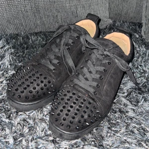 Svarta sneakers med nitar - Snygga svarta sneakers med nitar på ovansidan och en röd sula. Skorna har snörning och är tillverkade i mocka. Perfekta för att ge din outfit en edgy touch.