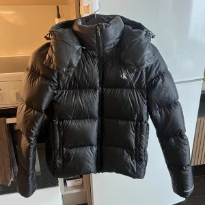 Svart dunjacka från Calvin Klein Jeans - Säljer en svart pufferjacka från Calvin Klein Jeans med huva och dragkedja. Jackan har en stilren design med CK-logga på bröstet och stor logga inuti. Perfekt för kyliga dagar. Inga anmärkningar förutom att den lilla ”upphängningsremsan” är av men annars i mycket bra skick. Dragkedjorna är hela och fina. Avtagbar luva. Inköptes på zalando för 2200kr för drygt 2 år sen och har mest bara hängt i garderoben - därav försäljning. Sista bilden är lånad från zalandos hemsida!