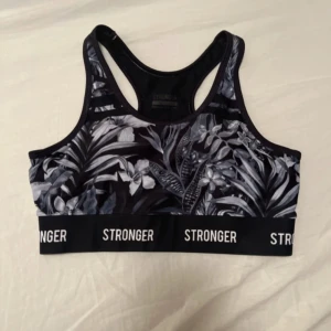 Svart blommig sporttopp från Stronger - Snygg svart sporttopp från Stronger med blommigt mönster i grått och vitt. Toppen har en elastisk kant med texten 'STRONGER' runtom. Perfekt för träning med sin bekväma passform och stilrena design.