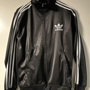 Svart träningsjacka från Adidas - Snygg svart träningsjacka från Adidas med klassiska vita ränder längs ärmarna och logga på bröstet. Jackan har dragkedja och ribbade muddar. Perfekt för sportiga tillfällen eller en avslappnad stil.