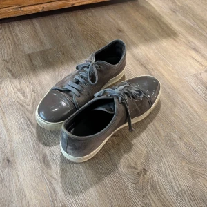 Grå sneakers från Lanvin - Snygga grå sneakers från Lanvin med en stilren design. Skorna har en glansig tå och snörning framtill. Perfekta för en avslappnad men elegant stil.