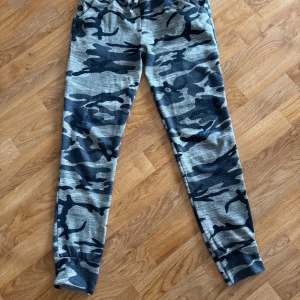 Camouflage mönstrade mjukisbyxor - Säljer ett par bekväma mjukisbyxor med grått och svart camouflagemönster. Byxorna har en snörning i midjan och muddar vid bensluten för en skön passform. Perfekta för en avslappnad stil.