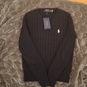 Svart stickad tröja från Ralph Lauren - Säljer en stilren svart stickad tröja från Ralph Lauren med v-ringning och det klassiska logotypbroderiet på bröstet. Tröjan har långa ärmar och ribbade muddar för en bekväm passform. Perfekt för kyligare dagar! Storleken ät SP vilket är en liten small.