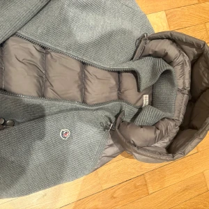 Grå stickad jacka med huva - Säljer en snygg grå stickad jacka från Moncler med huva och dragkedja. Jackan har långa ärmar och en quiltad insida för extra värme. Perfekt för kyliga dagar! Inget kvitto! Diskuterar pris vid snabb och smidig affär!!!