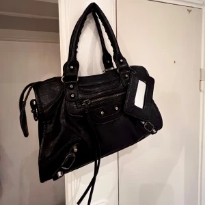 Svart axelväska med dragkedja - Snygg svart  skinnväska med dragkedja och nitar. Den har dubbla handtag och en avtagbar spegel. Avtagbara långa band tillkommer också ( se bild 3). Perfekt för att bära dina dagliga nödvändigheter med stil.