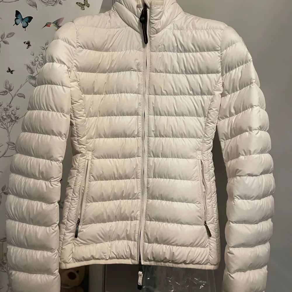 Säljer en snygg vit dunjacka från Parajumpers med dragkedja och hög krage. Jackan har en praktisk säkerhetsficka på insidan och en logotyp på ärmen. Perfekt för kyliga dagar.. Takit.