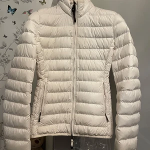 Vit dunjacka från Parajumpers - Säljer en snygg vit dunjacka från Parajumpers med dragkedja och hög krage. Jackan har en praktisk säkerhetsficka på insidan och en logotyp på ärmen. Perfekt för kyliga dagar.