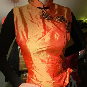 Orange sidenväst från Huế Silk - orange kinesisk/asiatisk väst från Huế Silk med broderade detaljer och ett par knappar framtill. vet ej om det är äkta siden eller inte! kan öppnas vid halsen. väldigt unik dragkedja baktill. tycker den är så snygg men har aldrig på mig den! stl S. 