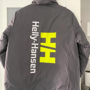 Hellu Hansen dunjacka - Vändbar dunjacka från Helly Hansen.  Grå m grönt tryck på ryggen el grå/svart m litet tryck på framsidan/arm. 