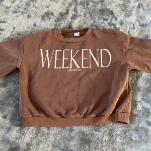 Säljer en brun sweatshirt från Lindex med texten 'WEEKEND forever' på framsidan. Tröjan är i storlek 146/152 och passar barn i åldern 10-12 år. Den är gjord av mjukt material och har långa ärmar samt ribbstickade muddar vid ärmslut och nederkant. Perfekt för en avslappnad helglook! 🧡