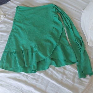 Grön omlottkjol från Vero Moda - Säljer en superfin grön omlottkjol från Vero Moda i storlek XXS. Kjolen har en volangdetalj längst ner och knyts i midjan för en perfekt passform. Den är gjord av ett lätt och luftigt material, perfekt för varma sommardagar. Använd ett fåtal gånger, så den är i nyskick. 