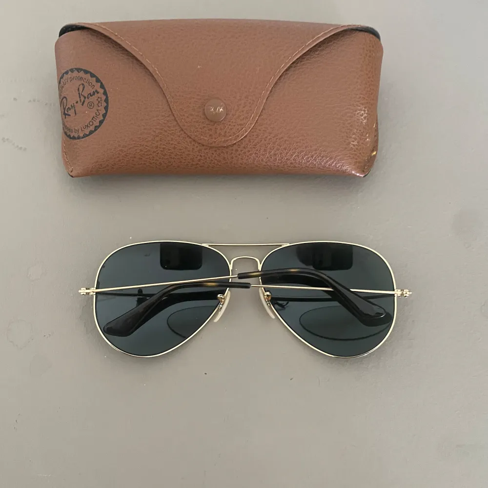 Säljer ett par klassiska Ray-Ban Aviator solglasögon med guldfärgad metallram och mörka linser. Glasögonen kommer med ett brunt läderfodral. Perfekta för soliga dagar och ger en tidlös look. Använda men i bra skick.. Accessoarer.